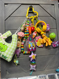 A colorful assortment of small charms, beads, and toy accessories hanging from a yellow carabiner on a dark gray wooden board. The collection includes a green and white checkered plush pouch with an apple patch that says 'MY DAY', pink cupcake and ice cream charms, rainbow beaded necklaces, a yellow My Little Pony figure, a green apple charm, and other pastel-colored decorative items.