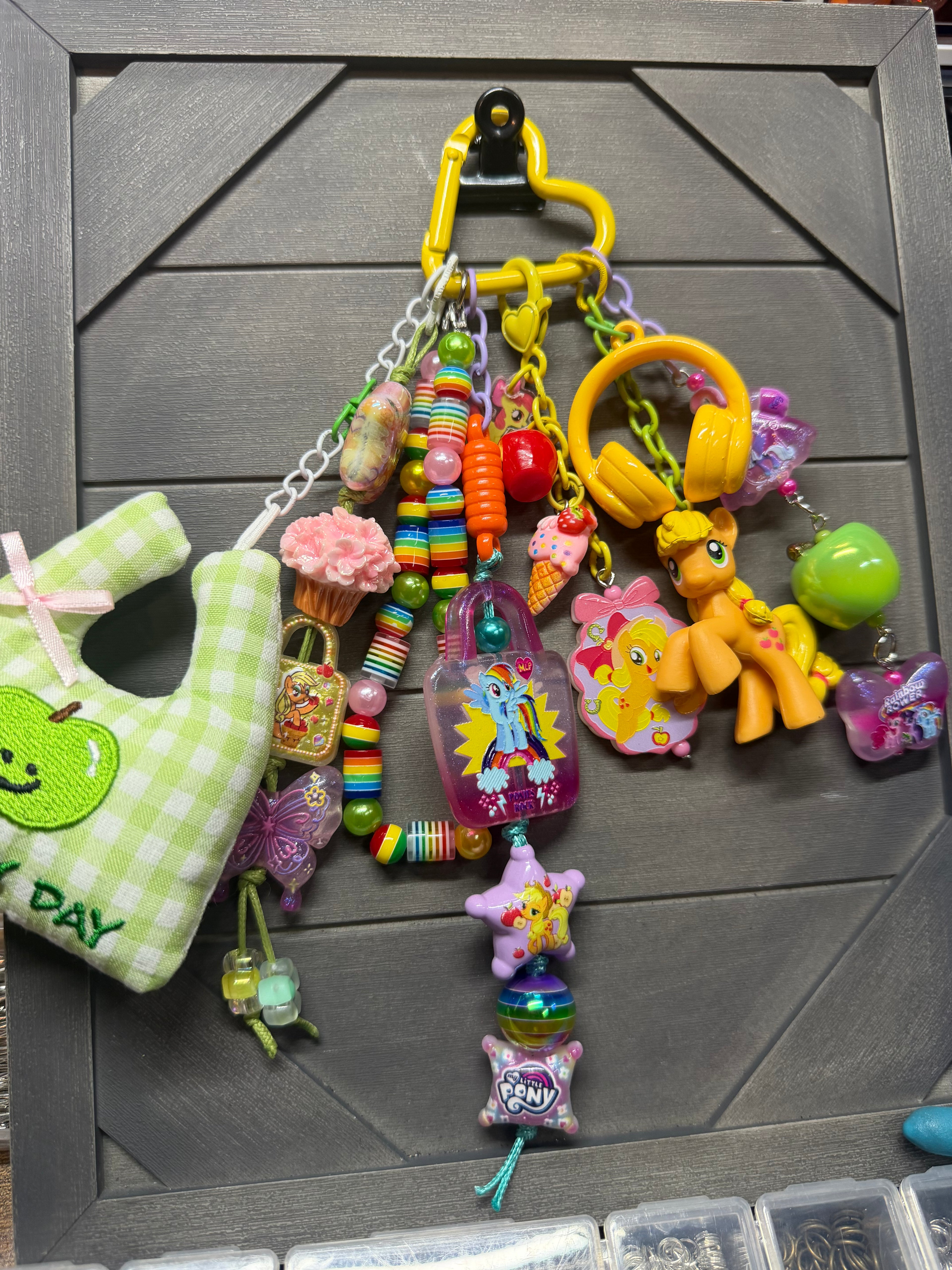 A colorful assortment of small charms, beads, and toy accessories hanging from a yellow carabiner on a dark gray wooden board. The collection includes a green and white checkered plush pouch with an apple patch that says 'MY DAY', pink cupcake and ice cream charms, rainbow beaded necklaces, a yellow My Little Pony figure, a green apple charm, and other pastel-colored decorative items.