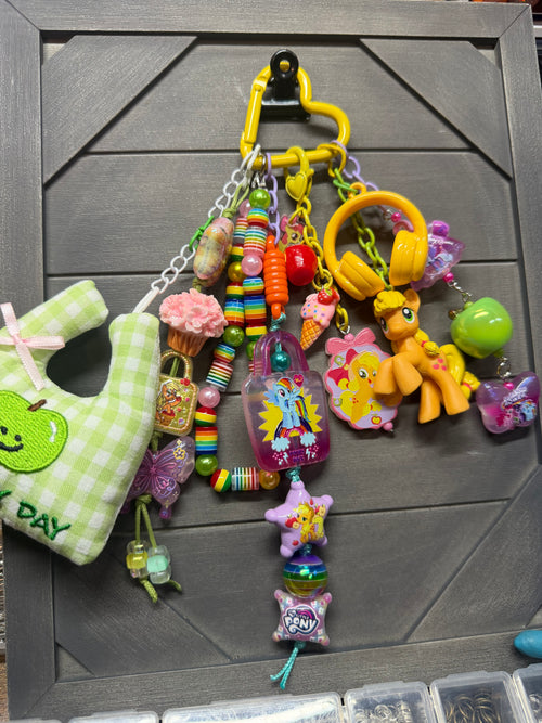 A colorful assortment of small charms, beads, and toy accessories hanging from a yellow carabiner on a dark gray wooden board. The collection includes a green and white checkered plush pouch with an apple patch that says 'MY DAY', pink cupcake and ice cream charms, rainbow beaded necklaces, a yellow My Little Pony figure, a green apple charm, and other pastel-colored decorative items.