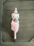 A pink Hello Kitty novelty pen with a fluffy pink pom-pom, a Hello Kitty character figure, and a small pink flower on top, photographed against a dark wood plank background.