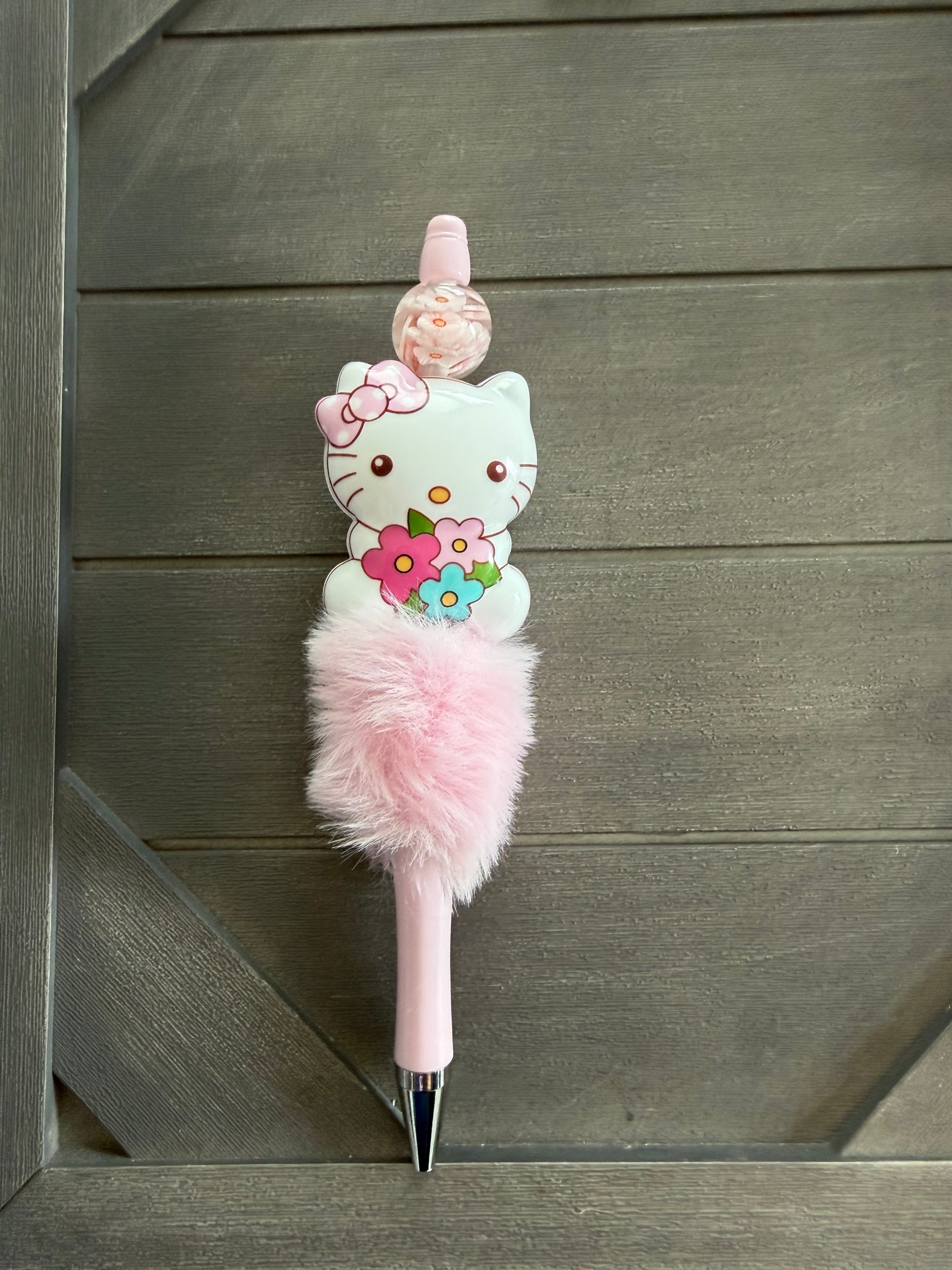A pink Hello Kitty novelty pen with a fluffy pink pom-pom, a Hello Kitty character figure, and a small pink flower on top, photographed against a dark wood plank background.