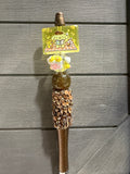 A decorative brown and gold pen with a cartoon charm featuring Pompompurin, a pink flower accent, and a rhinestone-embellished grip, set against a gray wood-paneled background.
