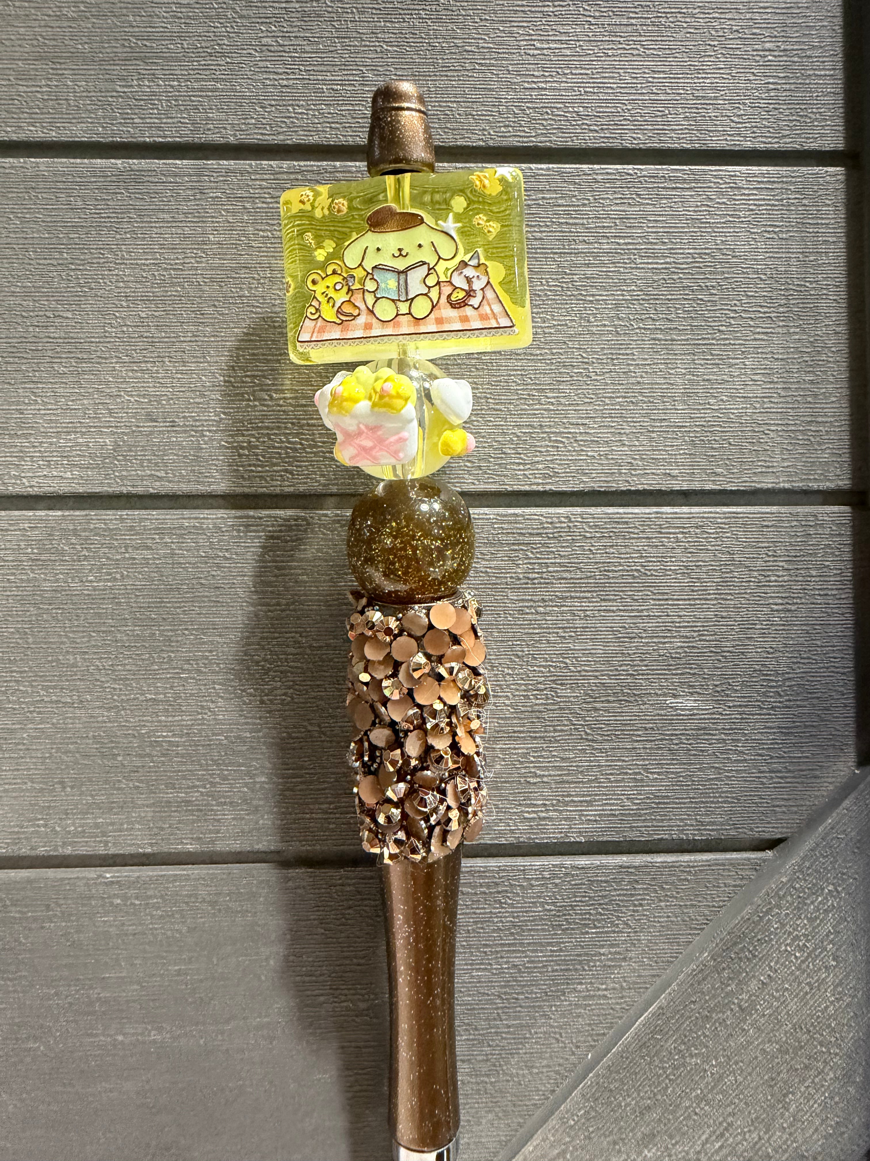 A decorative brown and gold pen with a cartoon charm featuring Pompompurin, a pink flower accent, and a rhinestone-embellished grip, set against a gray wood-paneled background.