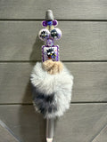 A decorative pen or stylus with a fluffy faux-fur body in gray, white, and black, topped with purple beads, owl-themed charms, and a small square card featuring a cartoon owl, set against a gray wood-paneled background.