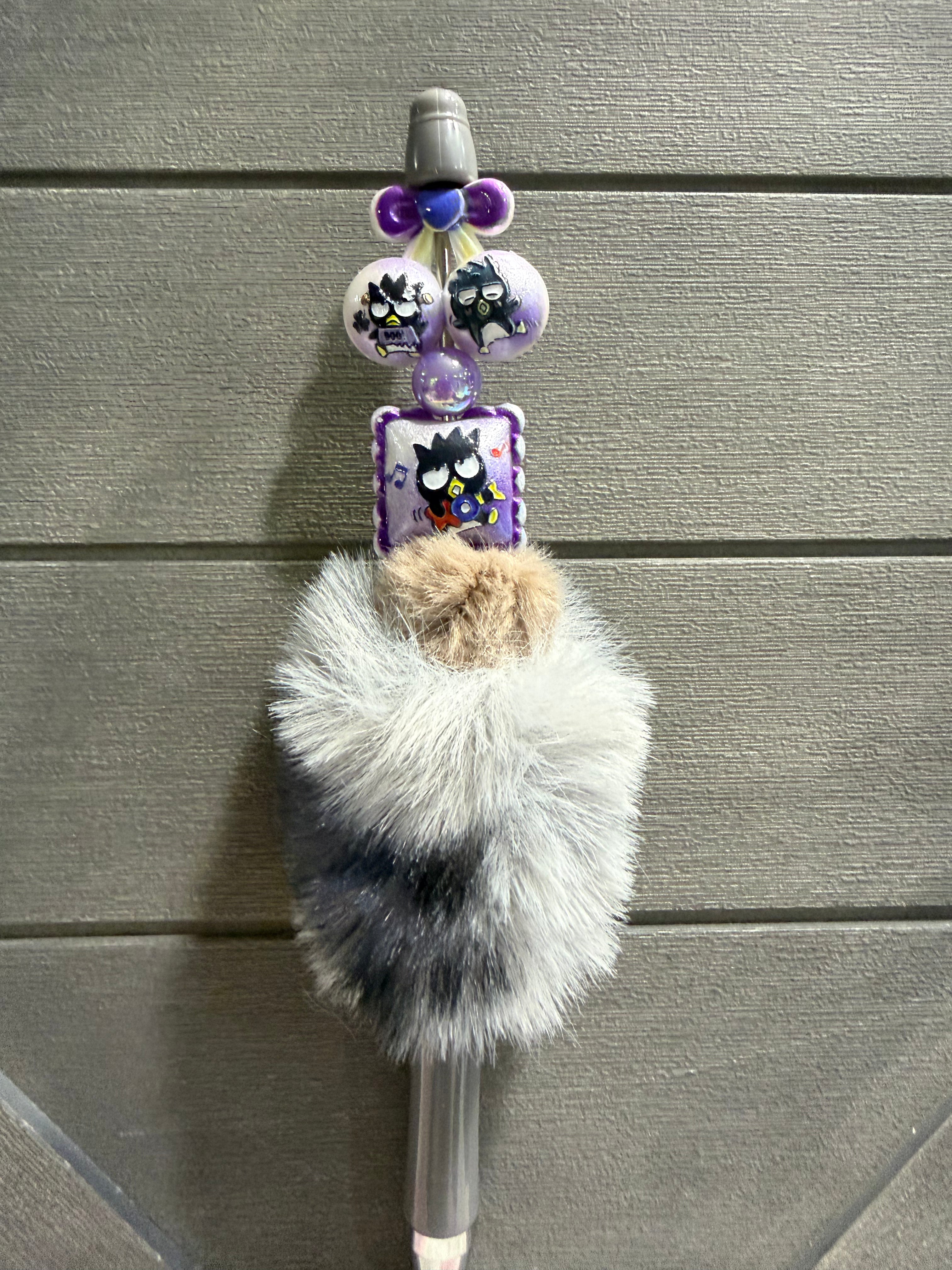 A decorative pen or stylus with a fluffy faux-fur body in gray, white, and black, topped with purple beads, owl-themed charms, and a small square card featuring a cartoon owl, set against a gray wood-paneled background.