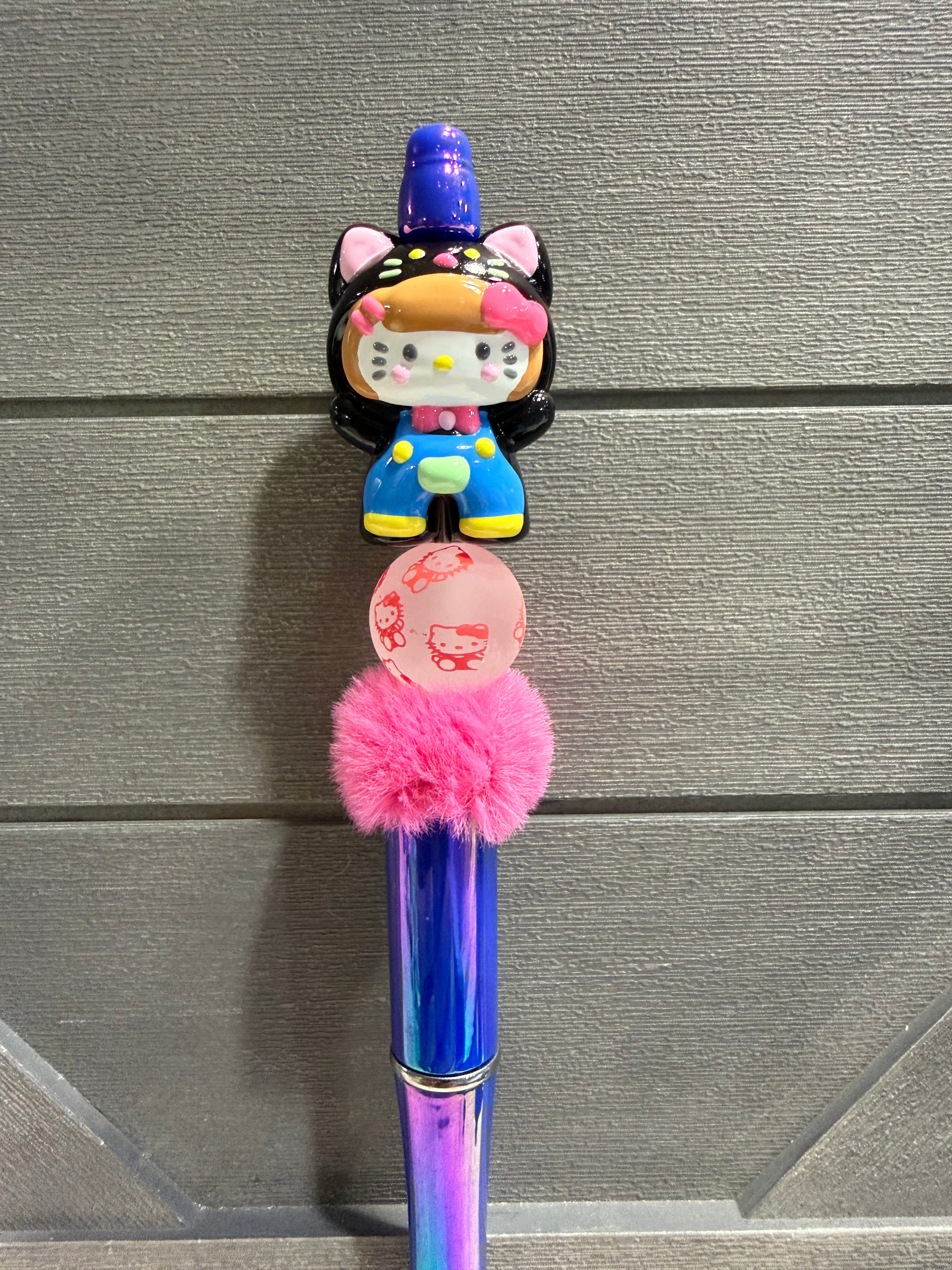 A decorative pen with a Hello Kitty character topper, a pink pom-pom, and a round pink charm, shown against a gray wood-textured background.