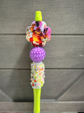 A novelty decorative pen or stylus with a bright lime green top and bottom, featuring a colorful floral charm, a textured purple bead, and a glittery decorative middle section, set against a gray wood-paneled background.