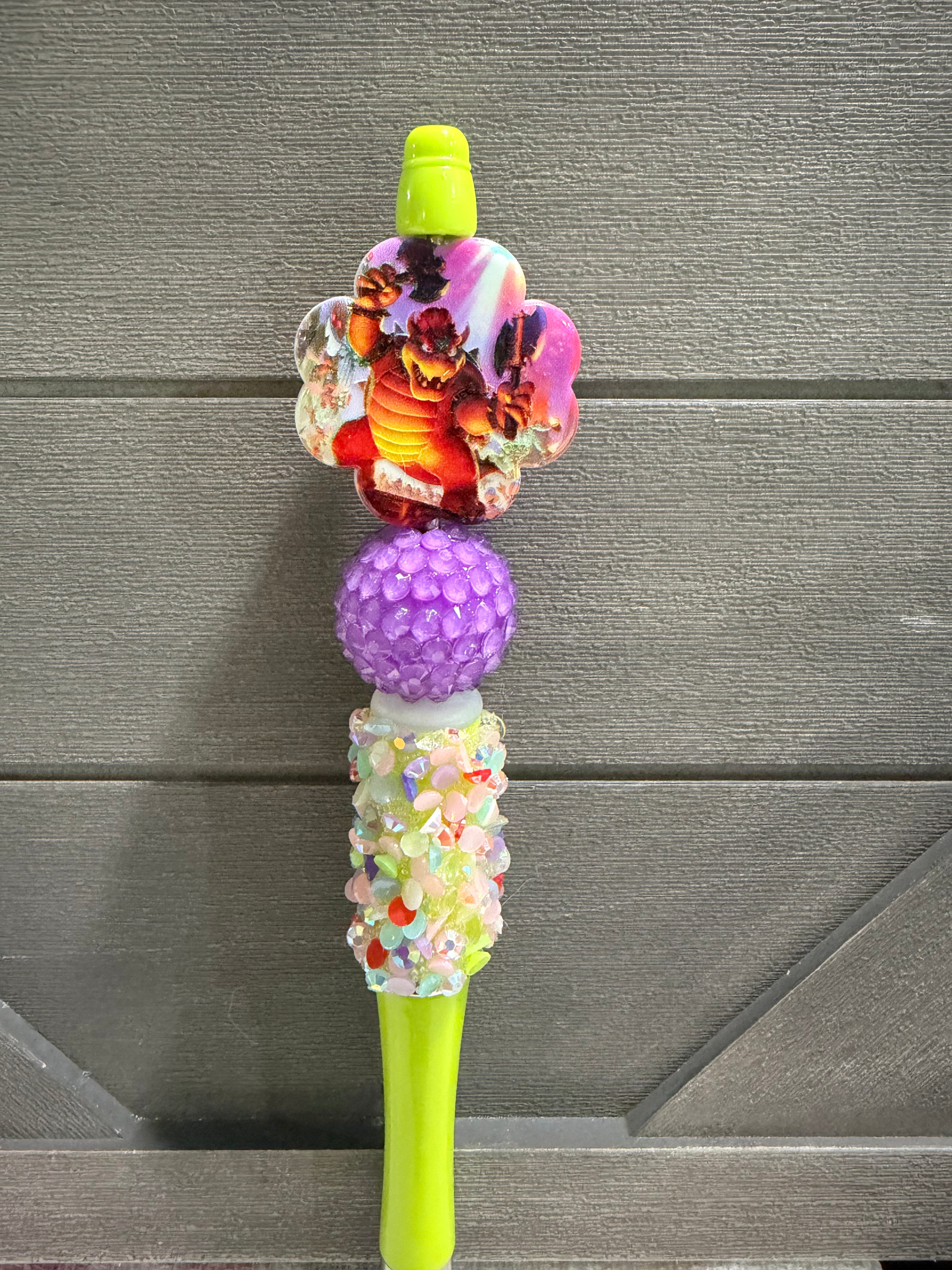 A novelty decorative pen or stylus with a bright lime green top and bottom, featuring a colorful floral charm, a textured purple bead, and a glittery decorative middle section, set against a gray wood-paneled background.