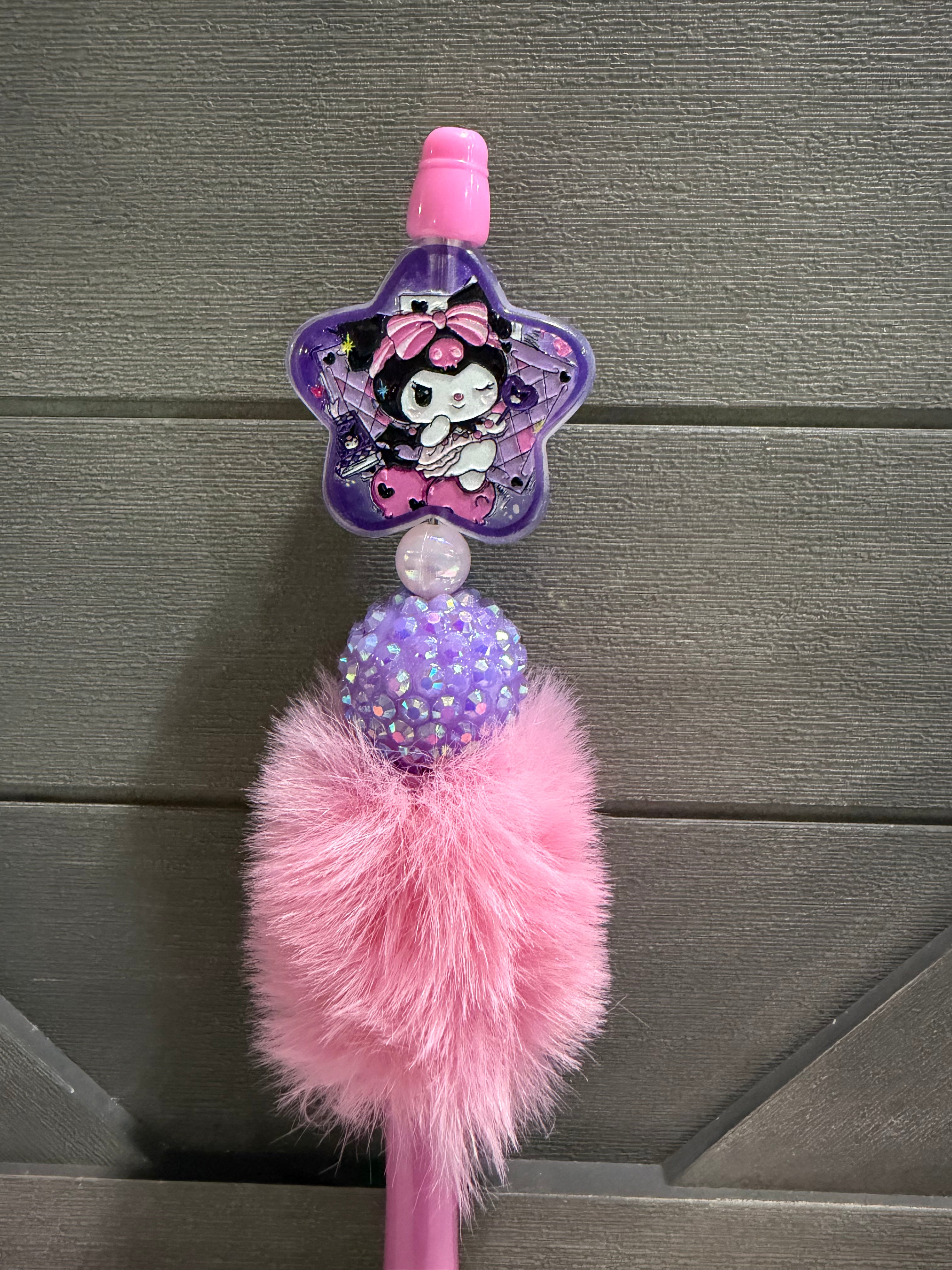 A pink and purple Kuromi-themed decorative pen or accessory with a star-shaped character charm, a glittery purple bead, and a fluffy pink pom-pom, shown against a gray wooden background.