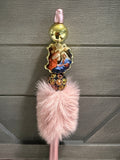 A pink religious rosary with a fluffy pink faux fur tassel, a gold-tone ball bead, a small religious medal depicting the Holy Family, and a rhinestone-encrusted spacer, shown against a gray wooden background.