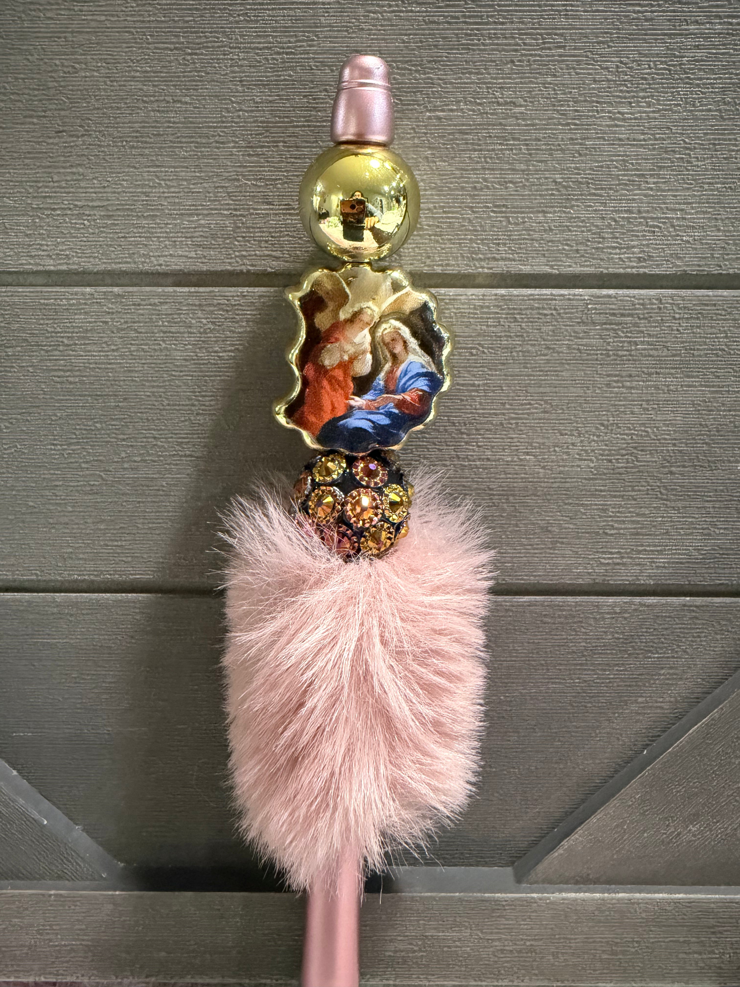 A pink religious rosary with a fluffy pink faux fur tassel, a gold-tone ball bead, a small religious medal depicting the Holy Family, and a rhinestone-encrusted spacer, shown against a gray wooden background.