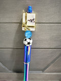 A decorative pen or novelty writing instrument with a holographic blue and purple barrel, a black-and-white soccer ball detail, a small gift box charm with gold trim, and a blue top cap, shown against a gray wood background.