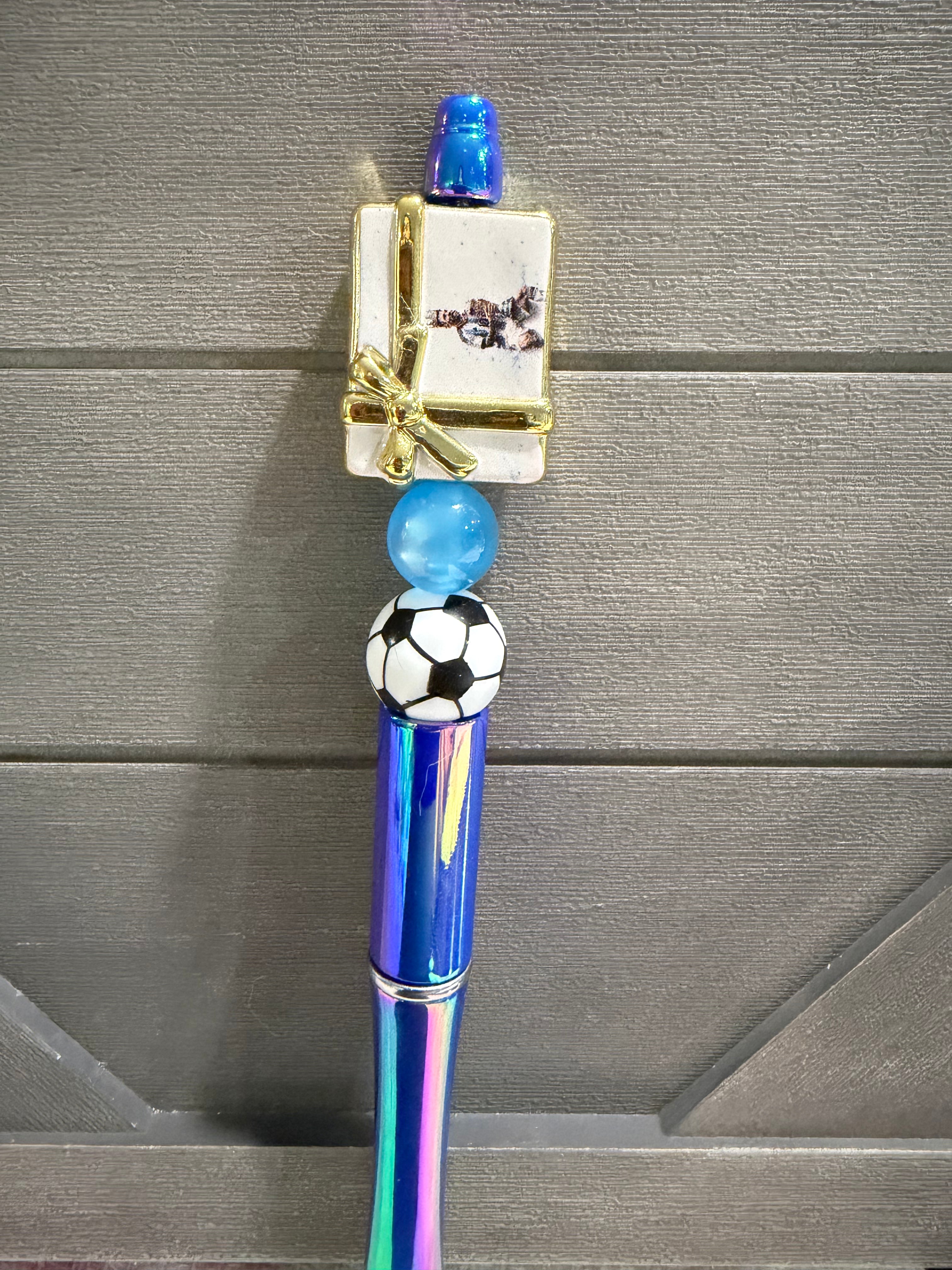 A decorative pen or novelty writing instrument with a holographic blue and purple barrel, a black-and-white soccer ball detail, a small gift box charm with gold trim, and a blue top cap, shown against a gray wood background.