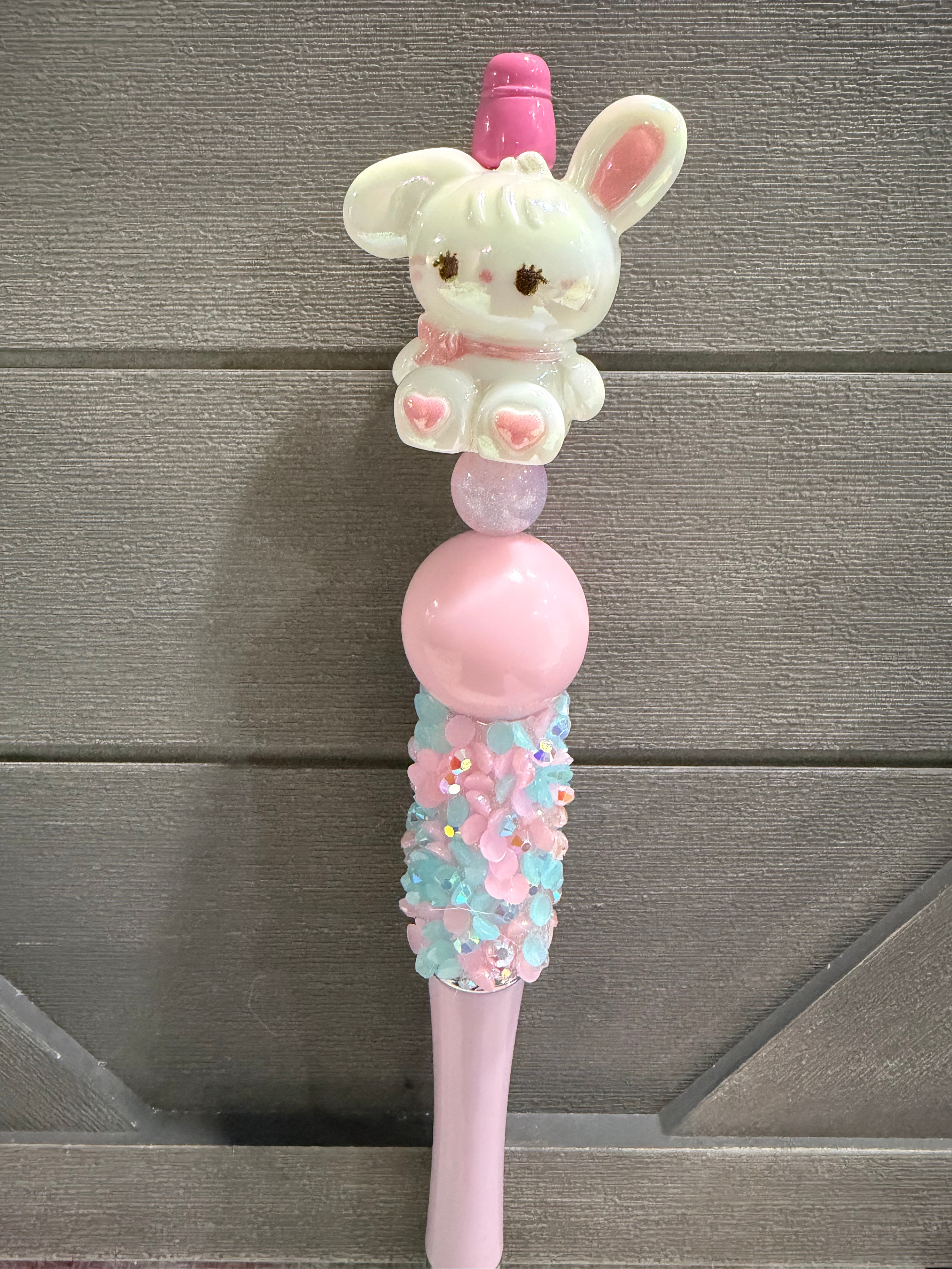 A pastel pink and white decorative pen or pencil featuring a small white bunny figure with pink ears and a pink top, a large pink oval center, and a floral textured lower section, shown against a gray wood plank background.