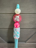 A decorative pen with a pastel blue handle, a pink and white cat-themed round ornament, and a textured top covered in pink and light blue rhinestones, set against a gray wood-paneled background.