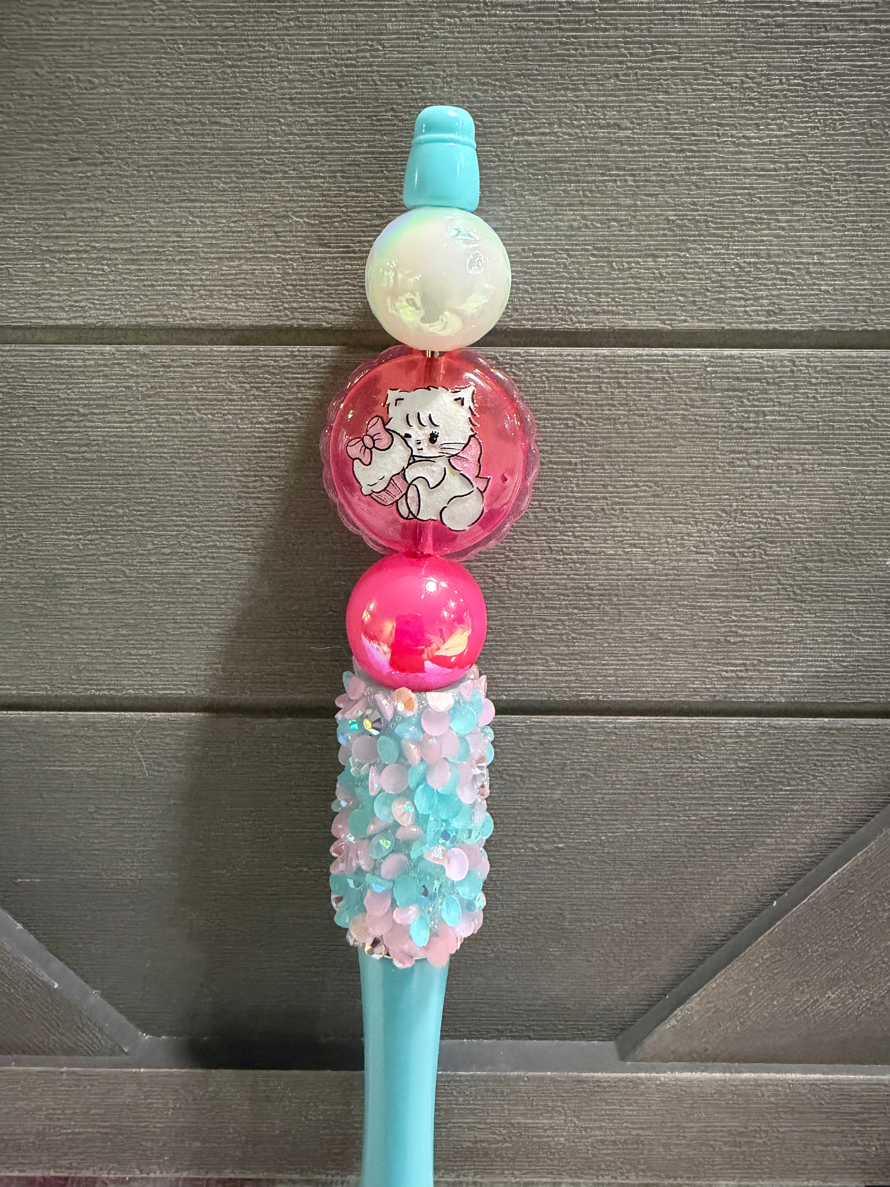 A decorative pen with a pastel blue handle, a pink and white cat-themed round ornament, and a textured top covered in pink and light blue rhinestones, set against a gray wood-paneled background.