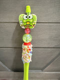 A green pen handle with a Kerrikyumon character face on the brush head, a red spacer bead, a clear bead filled with glitter, and a glittery cylindrical grip, set against a gray wood-paneled background.