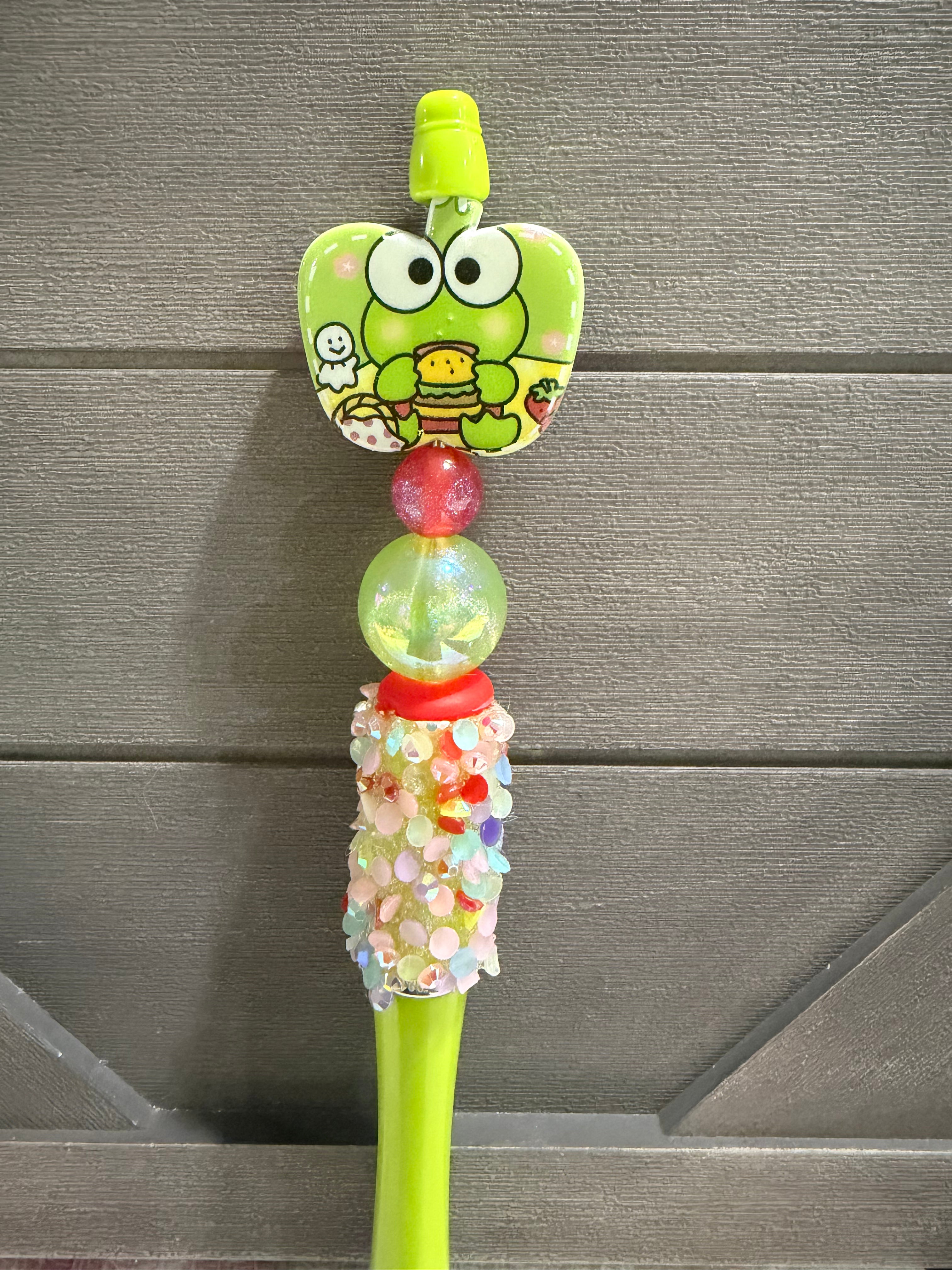 A green pen handle with a Kerrikyumon character face on the brush head, a red spacer bead, a clear bead filled with glitter, and a glittery cylindrical grip, set against a gray wood-paneled background.