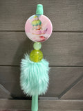 A mint green and pastel pink decorative pen with a round printed charm, a translucent yellow-green bead, and a fluffy mint green faux-fur tassel, shown against a dark wood-paneled background.