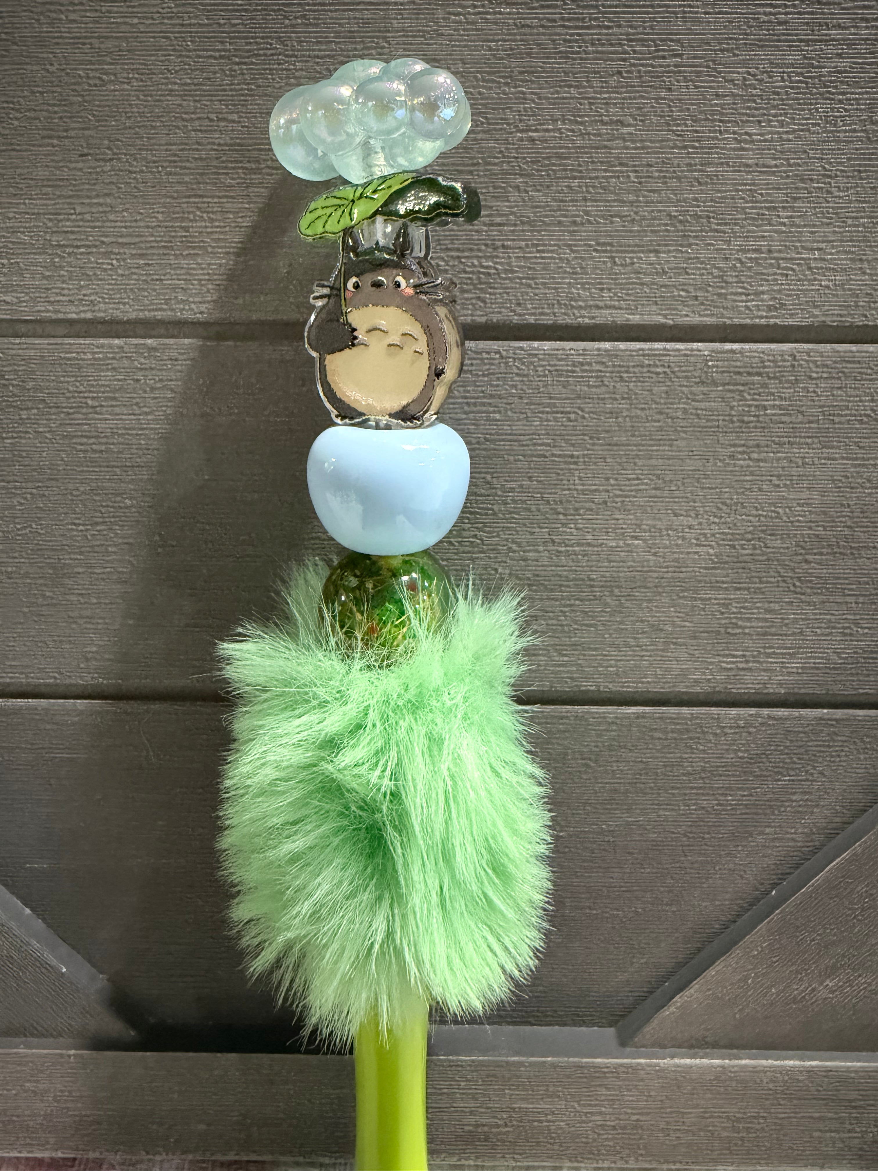 A decorative green-handled stick or tool with a fluffy mint green pom-pom, a translucent light blue round piece, a small character charm, green leaf and cloud details, and a metallic cap, shown against a dark wood plank background.