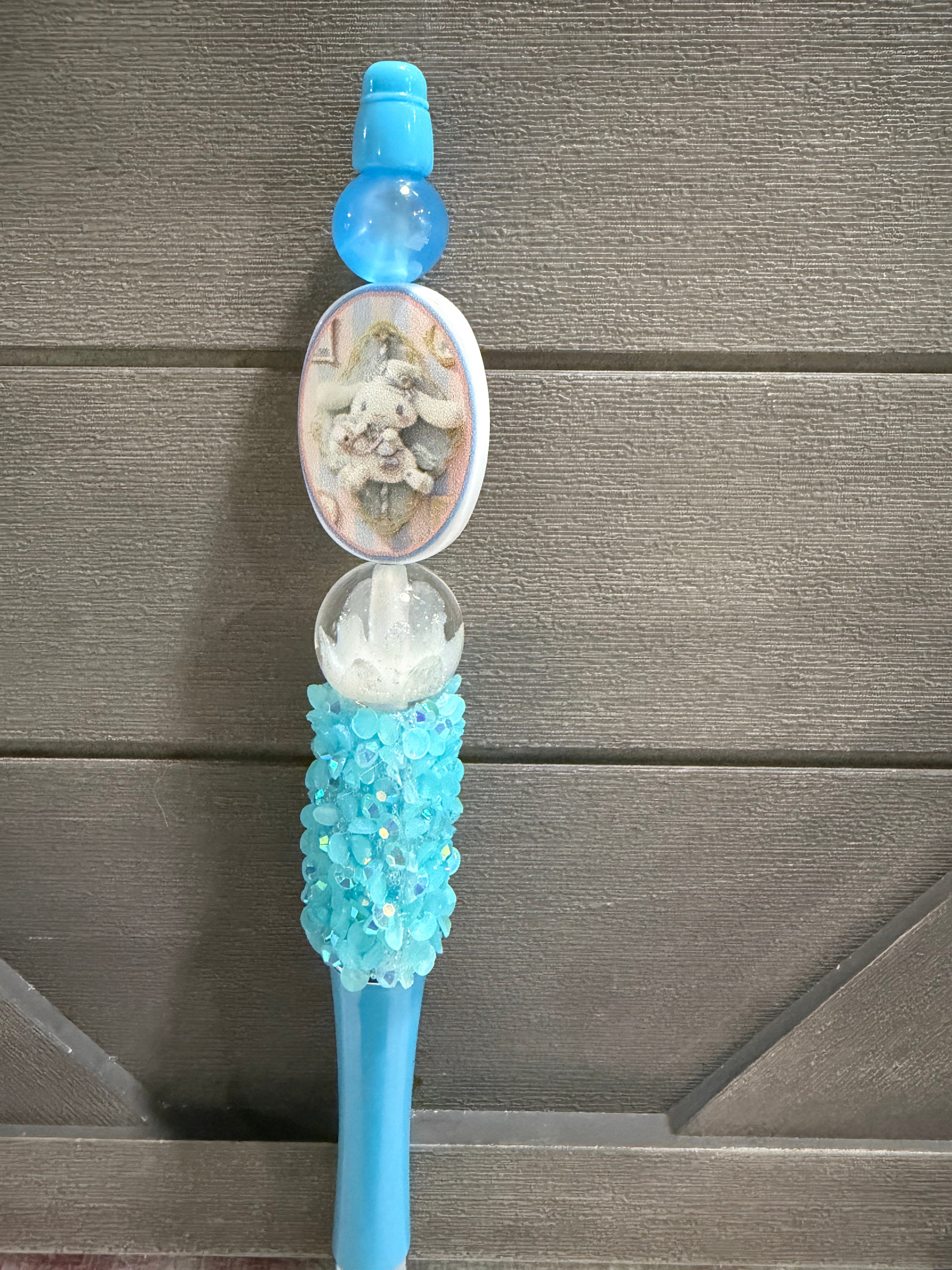 A decorative blue pen with a carved oval medallion, a white crystal-like top, and a textured turquoise handle, shown against a gray wood plank background.