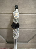 A decorative stylus or pen with a gray cartoon character topper, a clear round section filled with glitter, a black faux-fur accent, and a rhinestone-covered lower section, shown against a gray paneled background.
