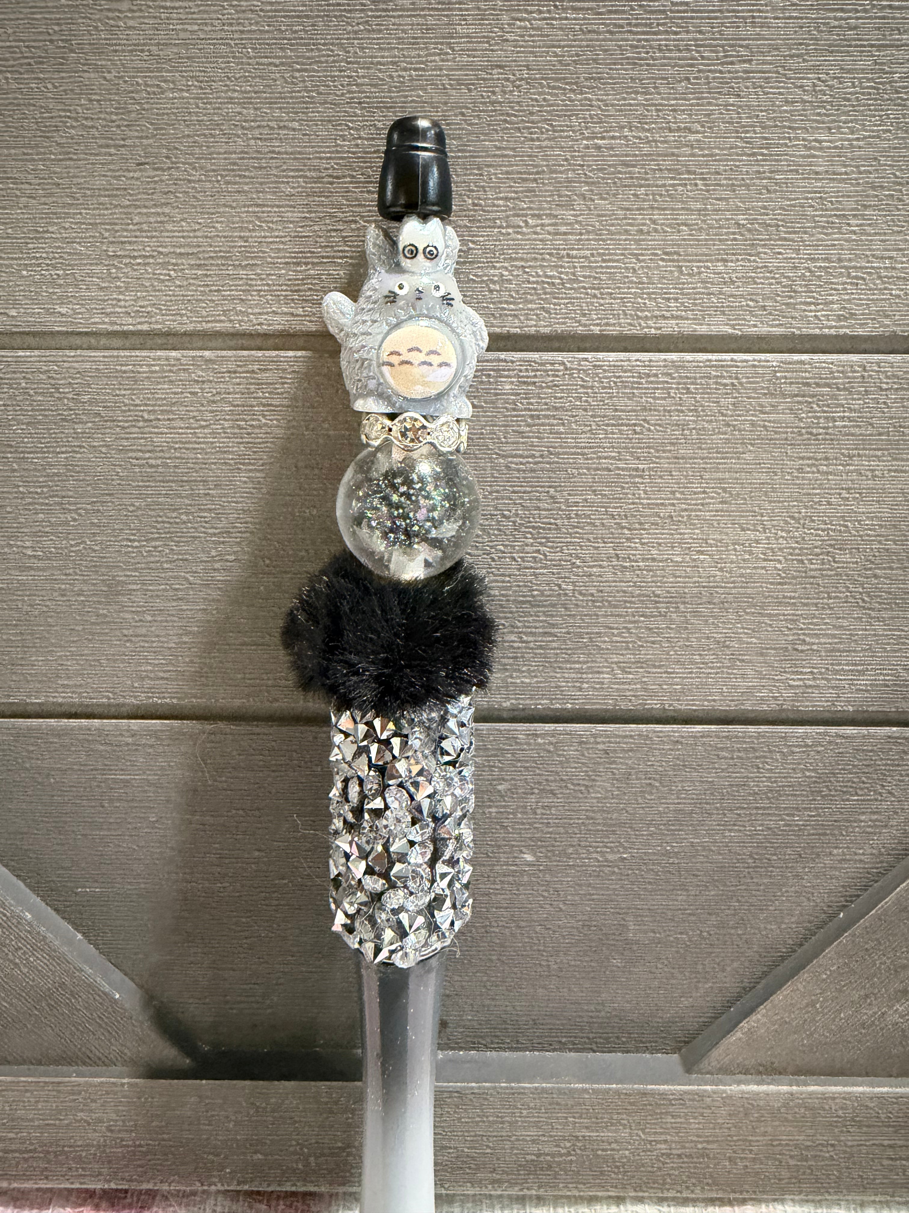 A decorative stylus or pen with a gray cartoon character topper, a clear round section filled with glitter, a black faux-fur accent, and a rhinestone-covered lower section, shown against a gray paneled background.