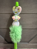 A pastel-colored beaded keychain or charm with a mint green fluffy pom-pom, a heart-shaped character bead, and a rhinestone-embellished round bead, shown against a dark wood background.