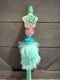 A mint green plush pen featuring a star-shaped Bulbasaur charm, a pink pom-pom, a blue bead, and a fluffy mint green faux fur tassel, shown against a dark wood background.