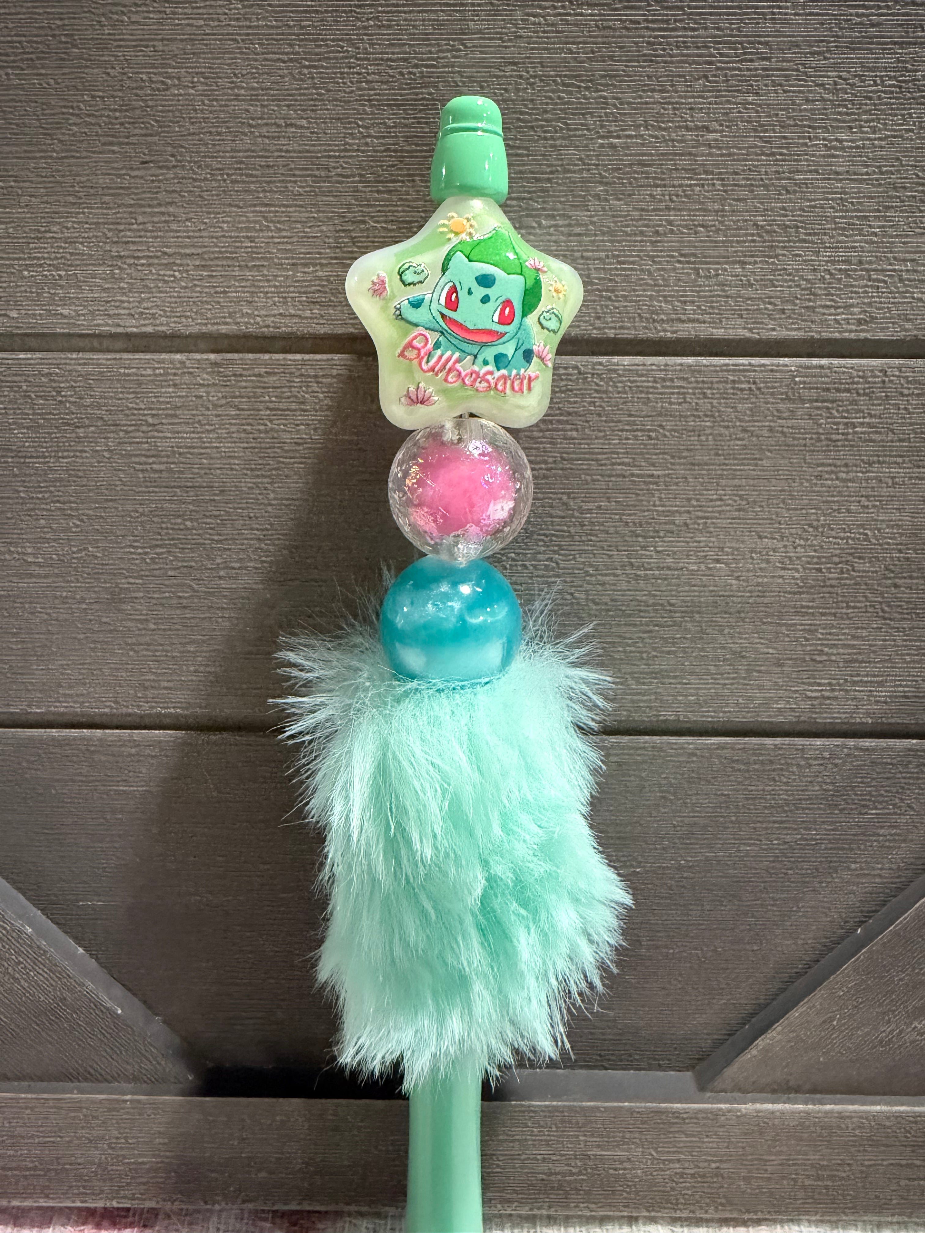 A mint green plush pen featuring a star-shaped Bulbasaur charm, a pink pom-pom, a blue bead, and a fluffy mint green faux fur tassel, shown against a dark wood background.
