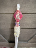 A pink and white decorative wand or stick with a Hello Kitty character charm, a pearl bead, a glittery pink sphere, and a white beaded handle, shown against a gray wood plank background.