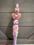 A pink Hello Kitty toothbrush with decorative beads, a heart-shaped charm, and a glittery handle, shown against a gray wooden background.