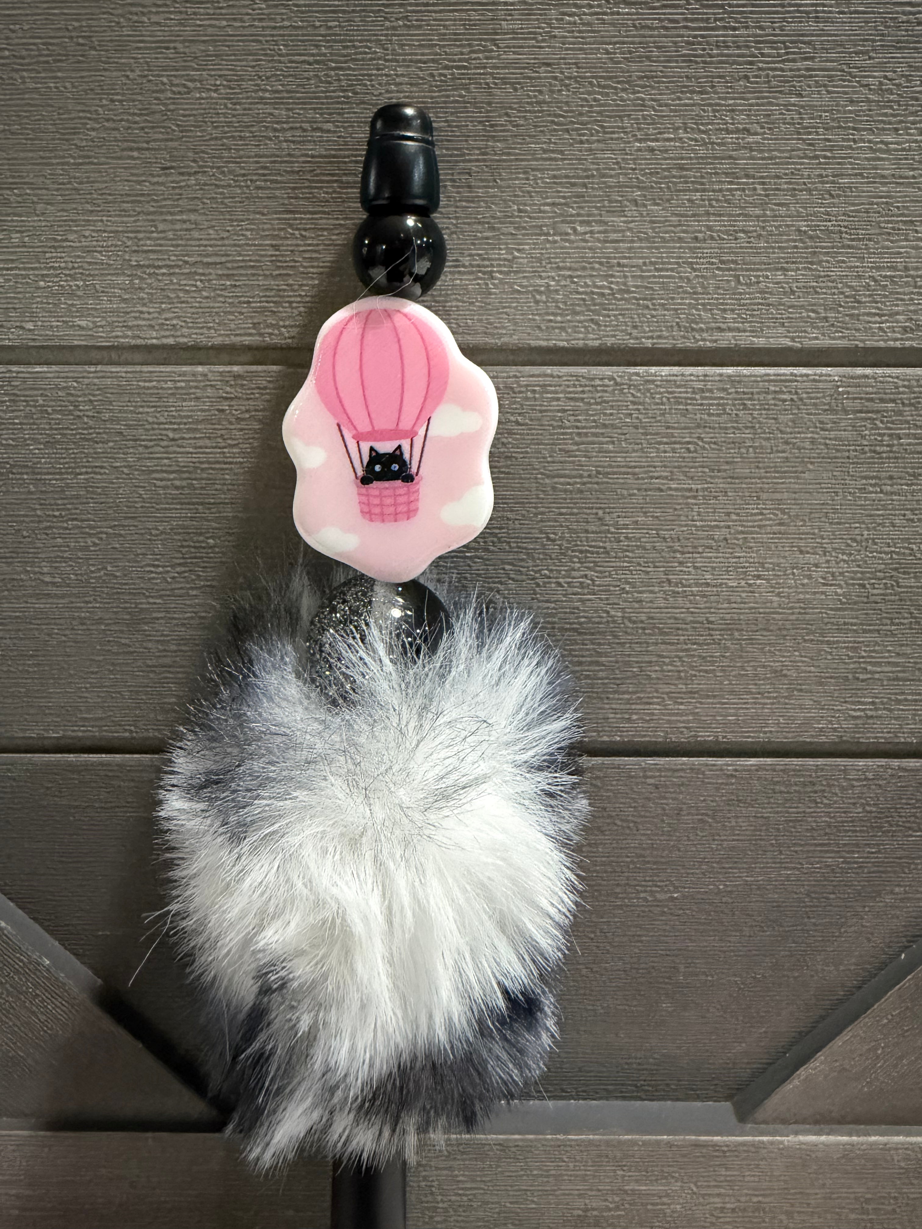 A black pen featuring a pink hot air balloon charm with a black cat in the basket, hanging from a fluffy white and gray faux fur pom-pom, set against a dark gray wood-paneled background.