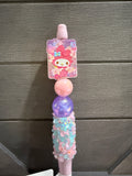 A pink and purple My Melody themed pen or stylus with a glittery pink top, a pink middle bead, a purple glitter middle bead, and a decorated lower handle, shown against a dark gray wood background.