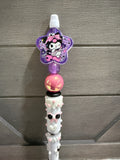 A close-up of a decorative pen or stylus with a purple star-shaped character charm, a pink glitter bead, and a white textured lower section, set against a gray wood-paneled background.