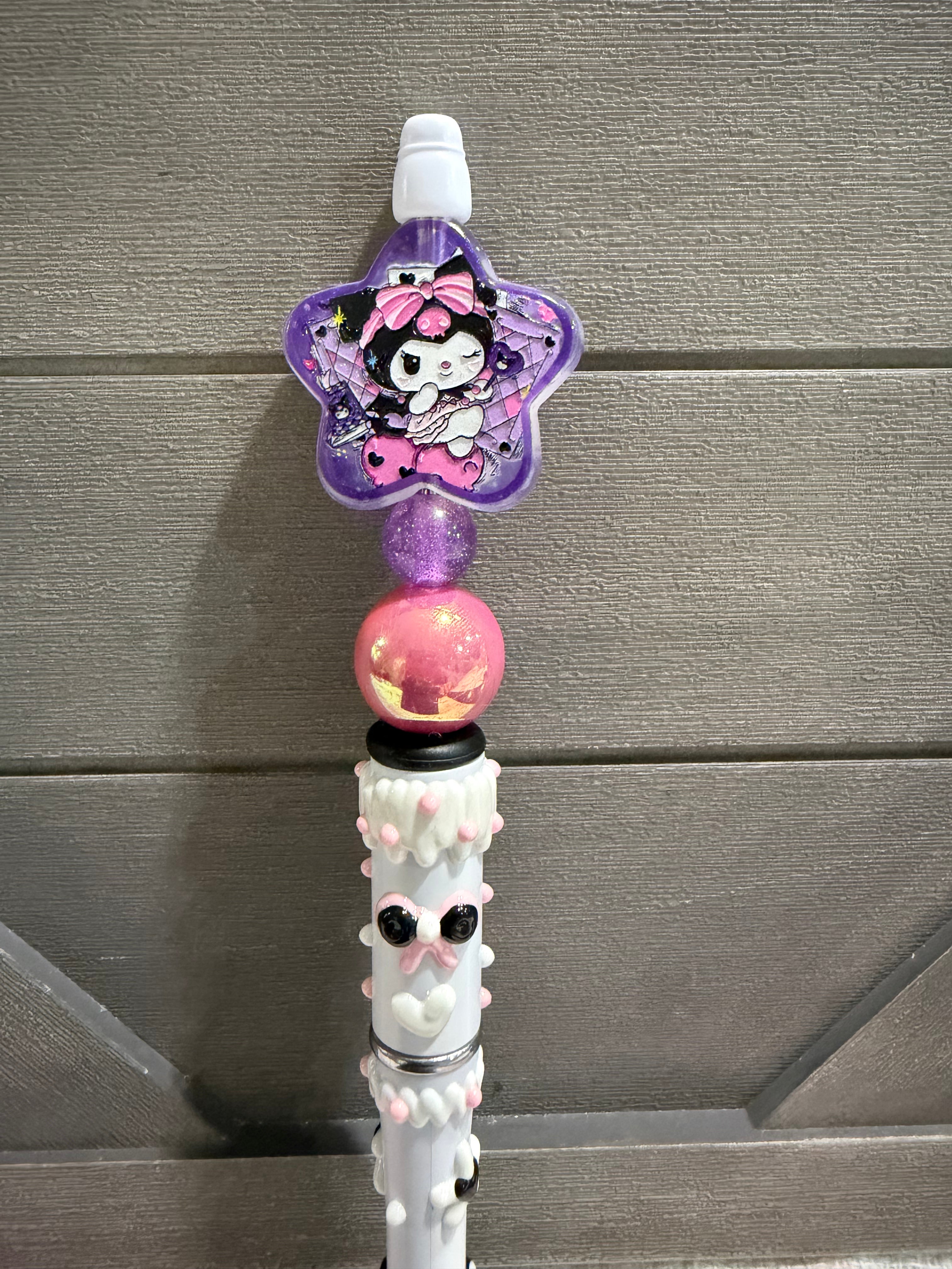 A close-up of a decorative pen or stylus with a purple star-shaped character charm, a pink glitter bead, and a white textured lower section, set against a gray wood-paneled background.