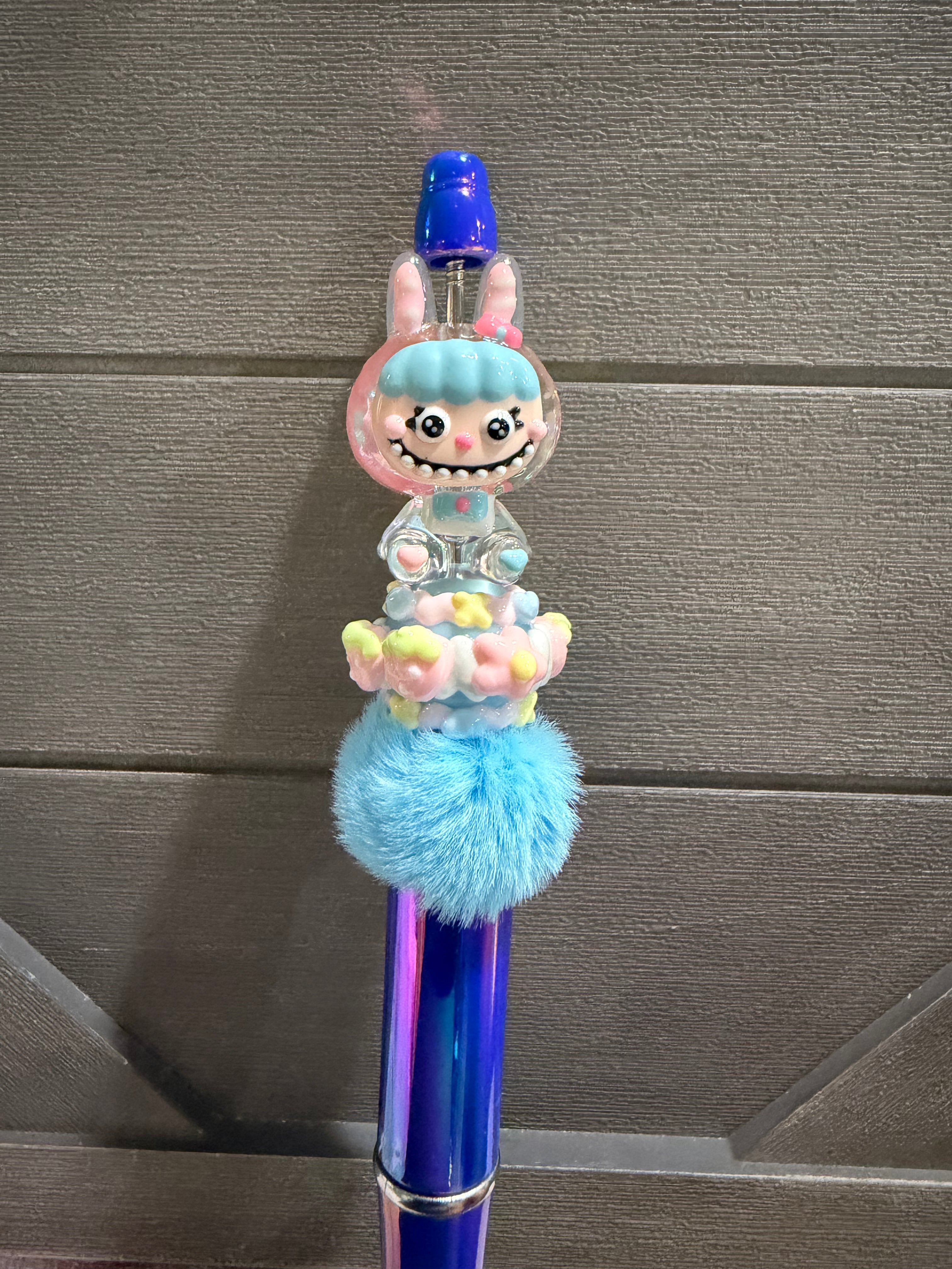 A decorative blue pen with a cartoon bunny character on top, surrounded by pastel flowers and a fluffy light blue pom-pom, set against a dark gray paneled background.