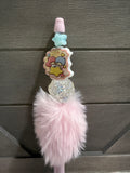 A pink and white decorative pen featuring a cartoon character sticker, a light blue star, a pink cap, a clear rhinestone section, and a fluffy pink faux-fur pom-pom, shown against a dark gray wood background.