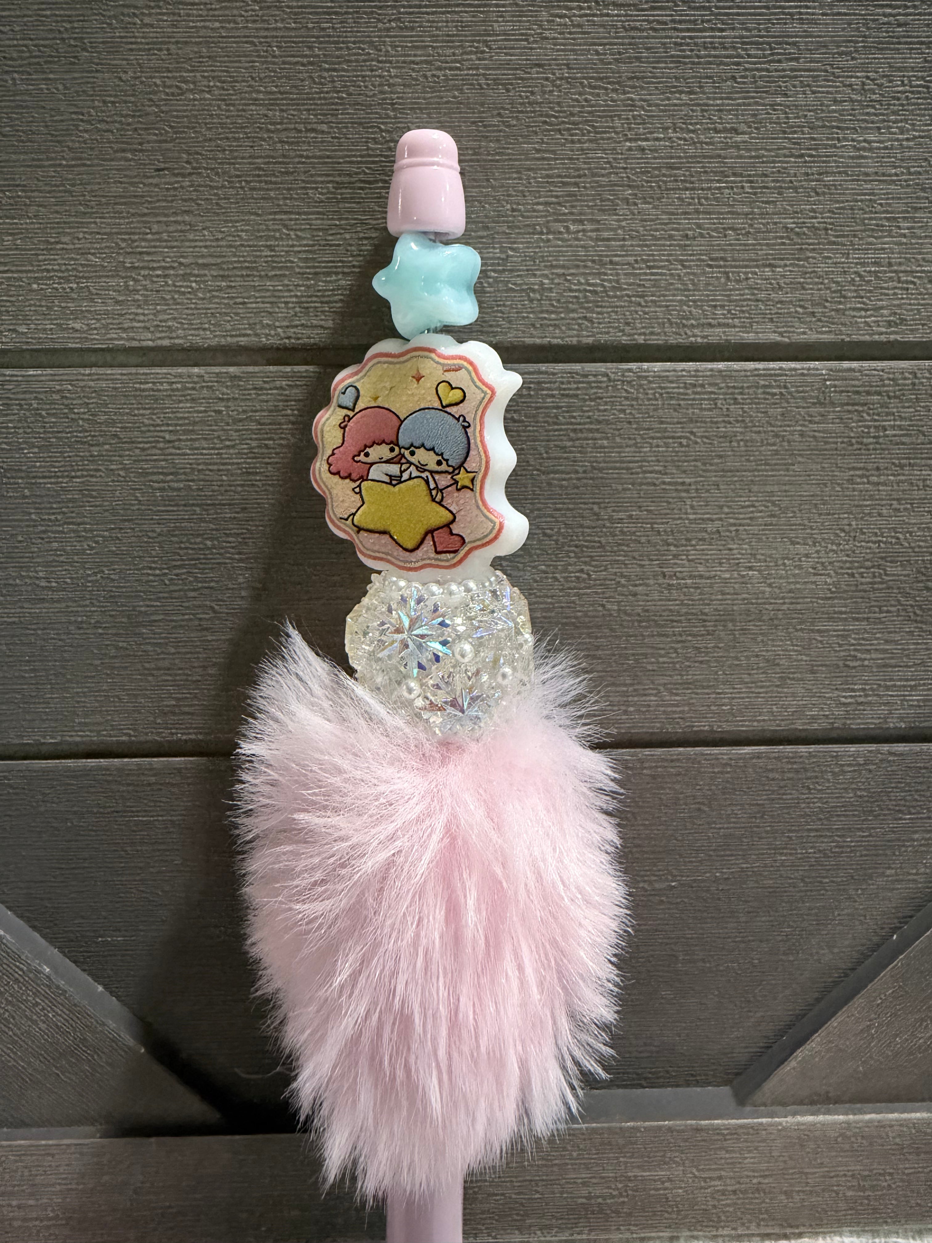 A pink and white decorative pen featuring a cartoon character sticker, a light blue star, a pink cap, a clear rhinestone section, and a fluffy pink faux-fur pom-pom, shown against a dark gray wood background.