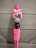 A pink decorative pen or stylus with anime character charms, rhinestone embellishments, and glossy beads, shown against a gray wood-paneled background.
