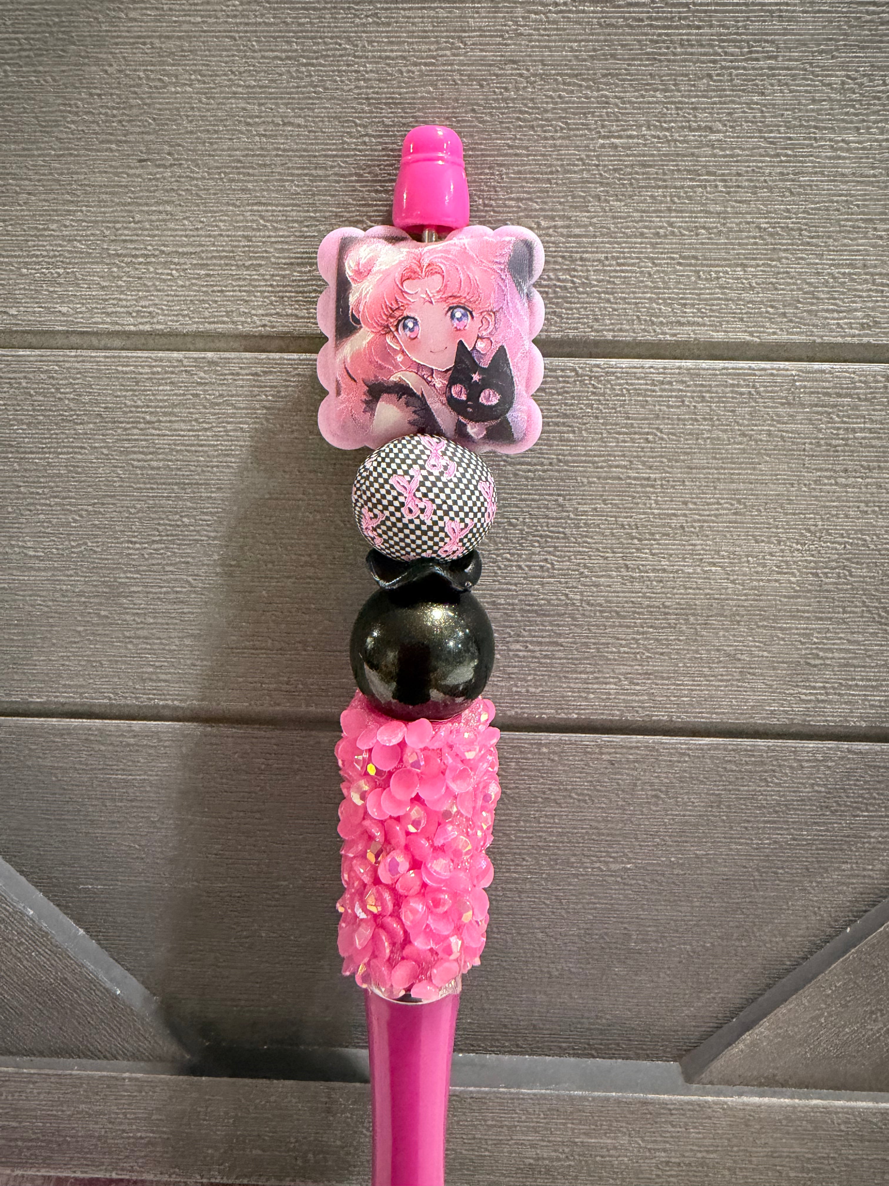 A pink decorative pen or stylus with anime character charms, rhinestone embellishments, and glossy beads, shown against a gray wood-paneled background.