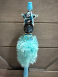 A light blue and turquoise decorative pen or novelty item with a fluffy faux-fur pom-pom, a dark starry bead, a small illustrated figure, and a blue top cap, photographed against a brown paneled background.