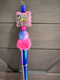 A decorative pen with a holographic blue and pink handle, a pink pom-pom, a purple bead, and a cartoon character charm, shown against a dark gray wooden background.