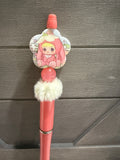 A pink decorative pen or stylus with a cartoon character charm, a fluffy white pom-pom, and a metallic ring, shown against a dark gray wood-paneled background.