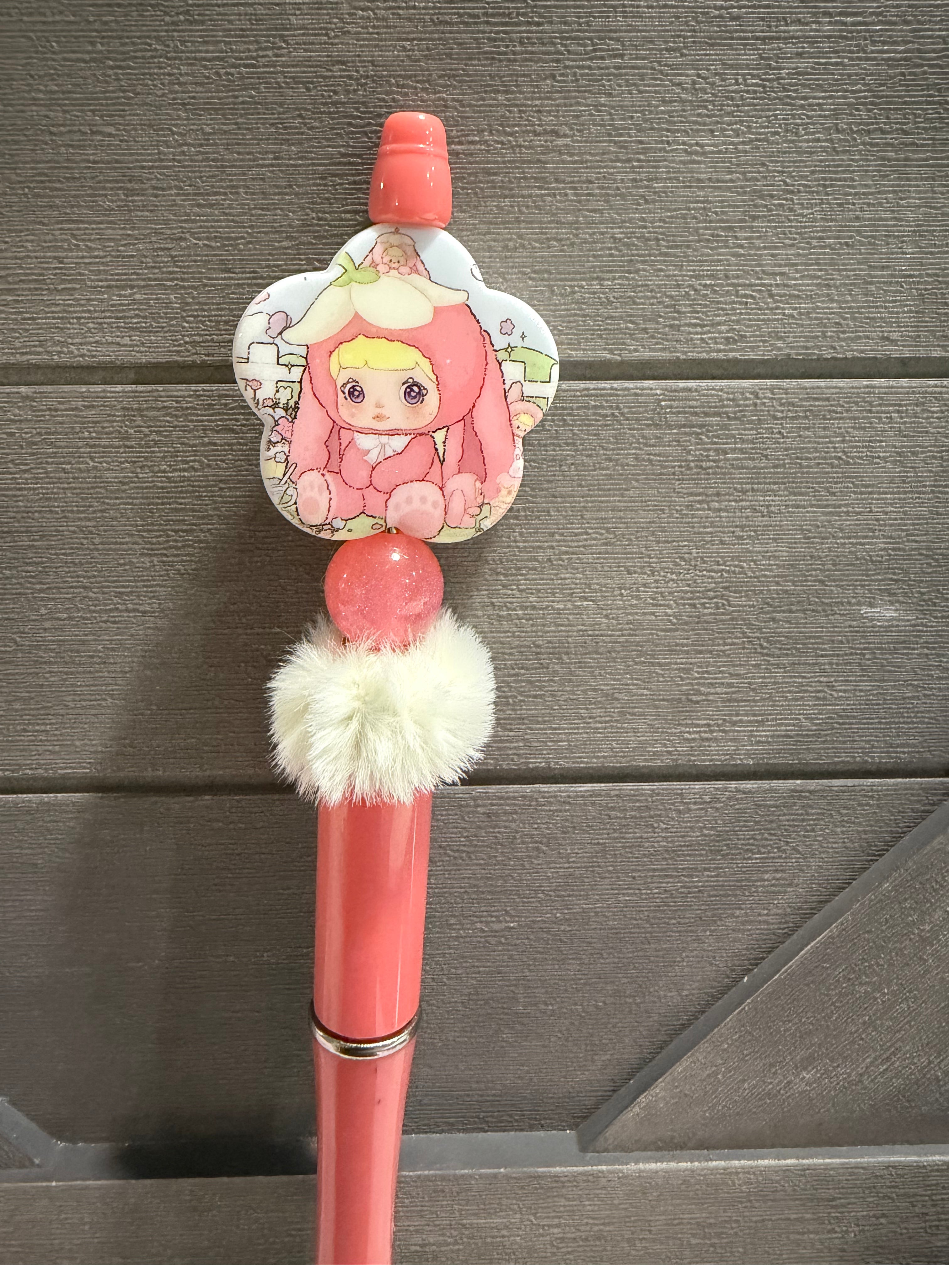 A pink decorative pen or stylus with a cartoon character charm, a fluffy white pom-pom, and a metallic ring, shown against a dark gray wood-paneled background.