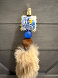 A decorative pen with a small square photo frame showing soccer players, a gold bell at the top, a blue bead, a brown wooden bead, and a large beige faux-fur tassel, hanging against a dark gray paneled background.