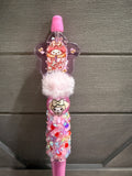 A pink decorative pen or stylus with a star-shaped clear top containing a cartoon character, a fluffy white pom-pom, and a glittery lower section with multicolored sequins, shown against a gray wood background.