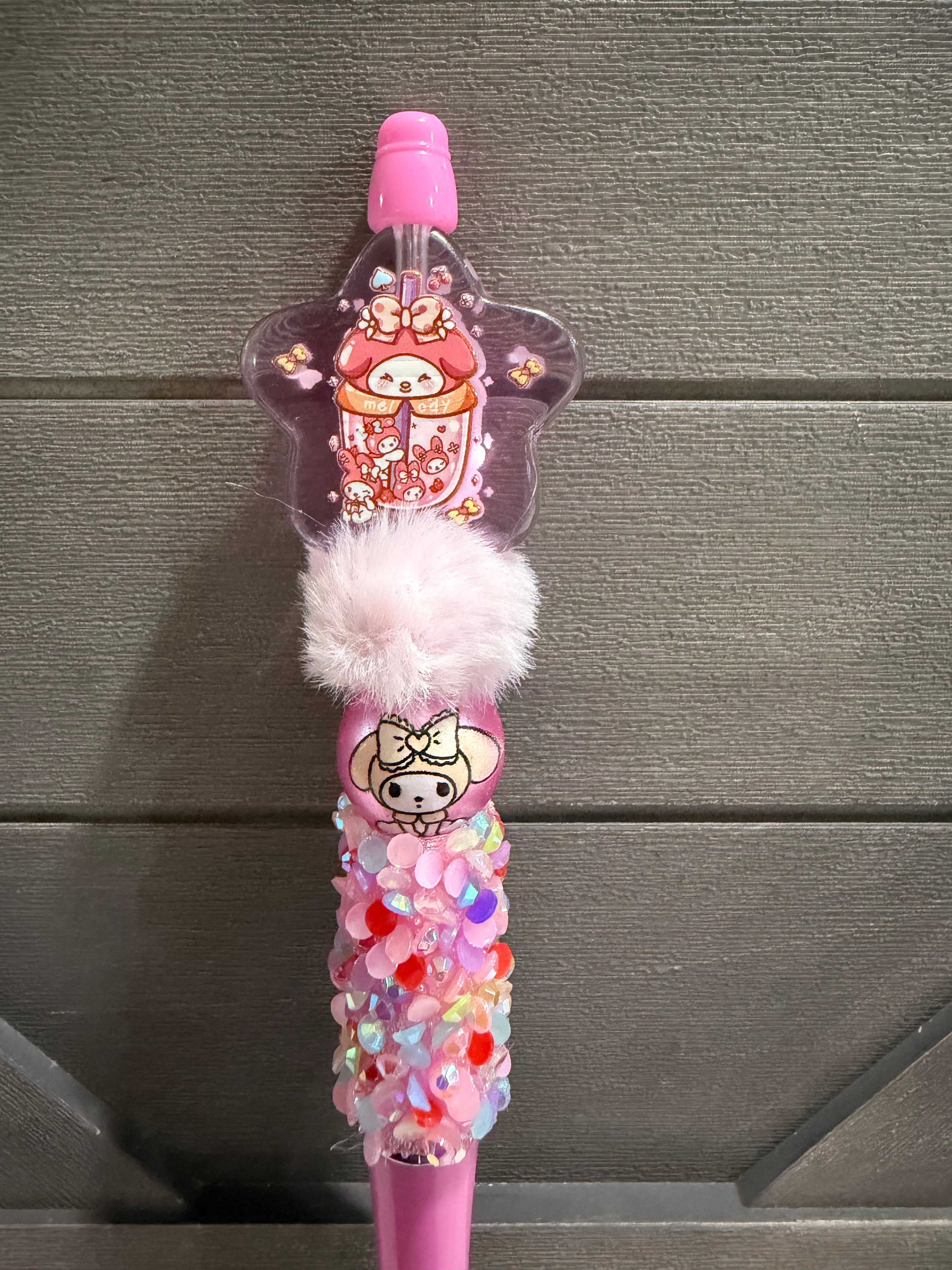 A pink decorative pen or stylus with a star-shaped clear top containing a cartoon character, a fluffy white pom-pom, and a glittery lower section with multicolored sequins, shown against a gray wood background.