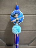 A blue decorative pen or stylus with a character illustration on a scalloped shell-shaped charm, a fluffy light blue pom-pom, and a shiny blue barrel, shown against a gray wood-textured background.