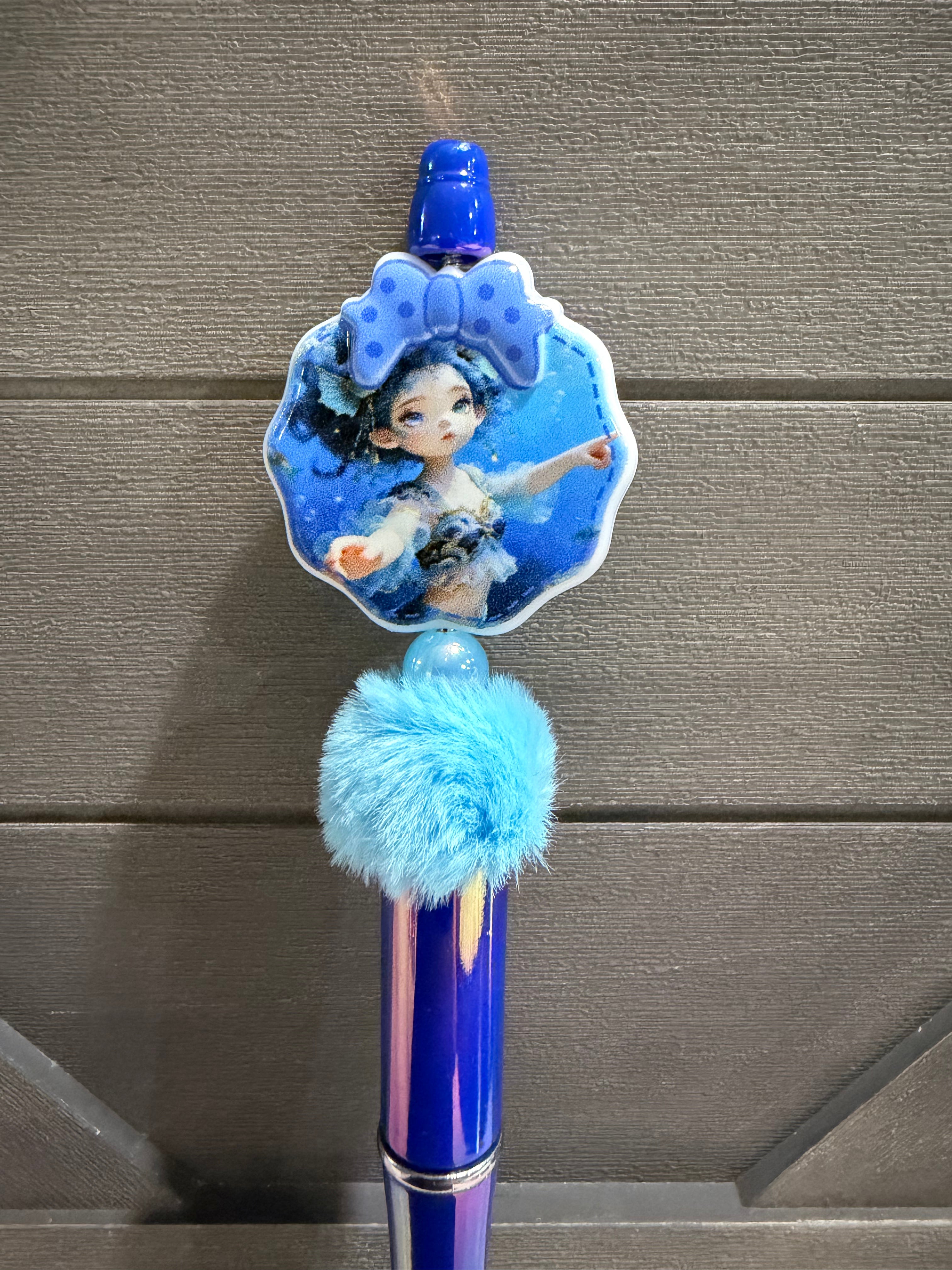 A blue decorative pen or stylus with a character illustration on a scalloped shell-shaped charm, a fluffy light blue pom-pom, and a shiny blue barrel, shown against a gray wood-textured background.